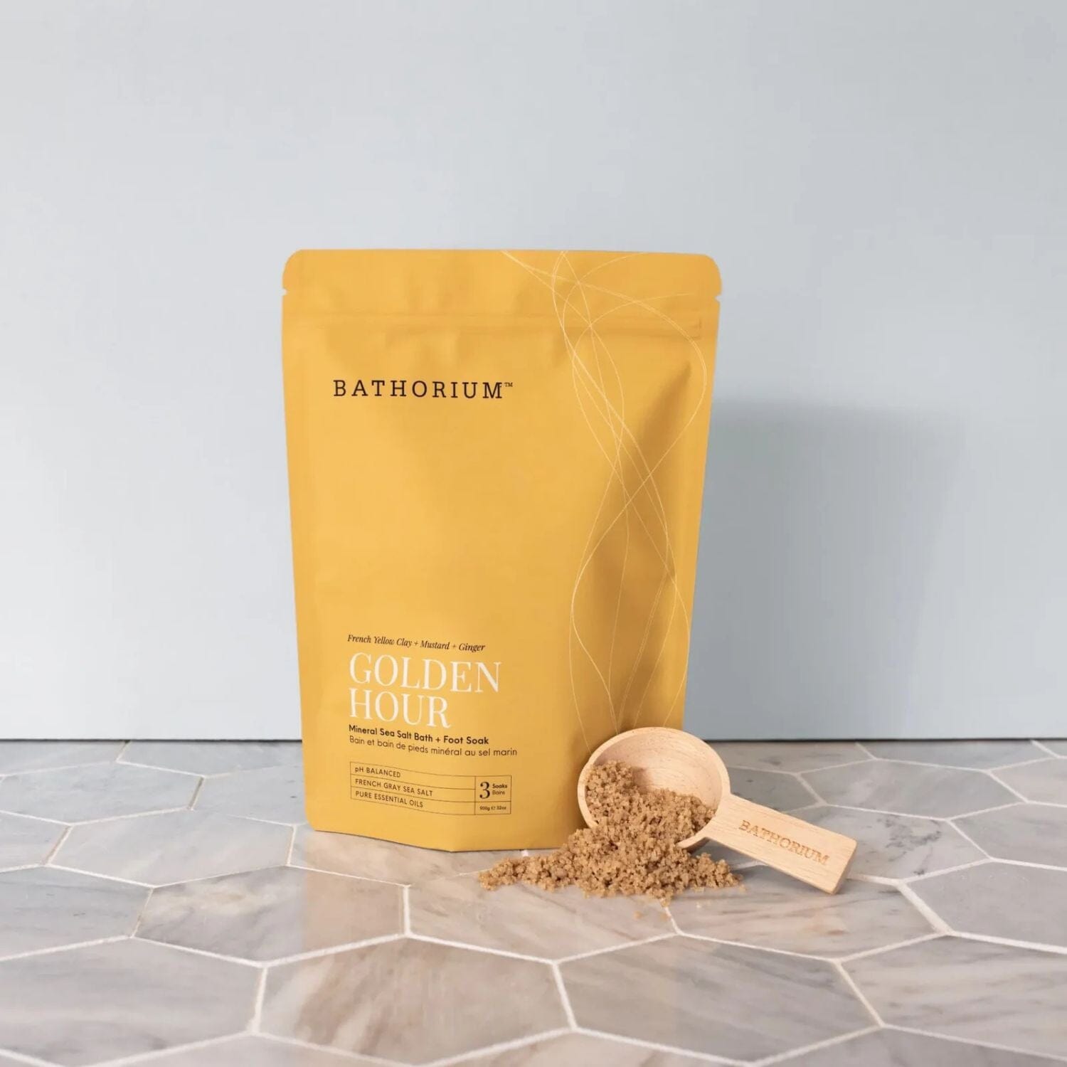 A yellow Bathorium Golden Hour almond sea salt scrub pouch stands on a gray marble surface with a small wooden scoop filled with the scrub beside it.