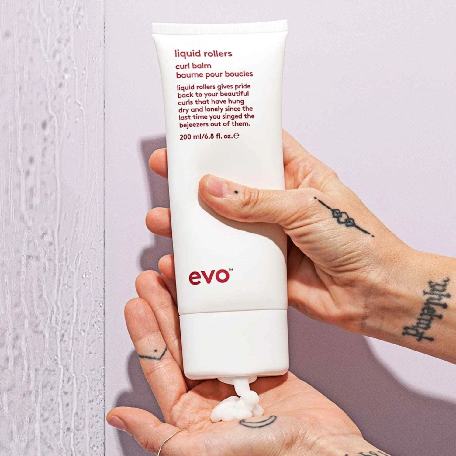 A hand holds a white squeeze tube of Evo Liquid Rollers curl balm, with some white product squeezed onto a palm below. The background is a split design with a light textured gray left side and a pale lavender/pink right side.