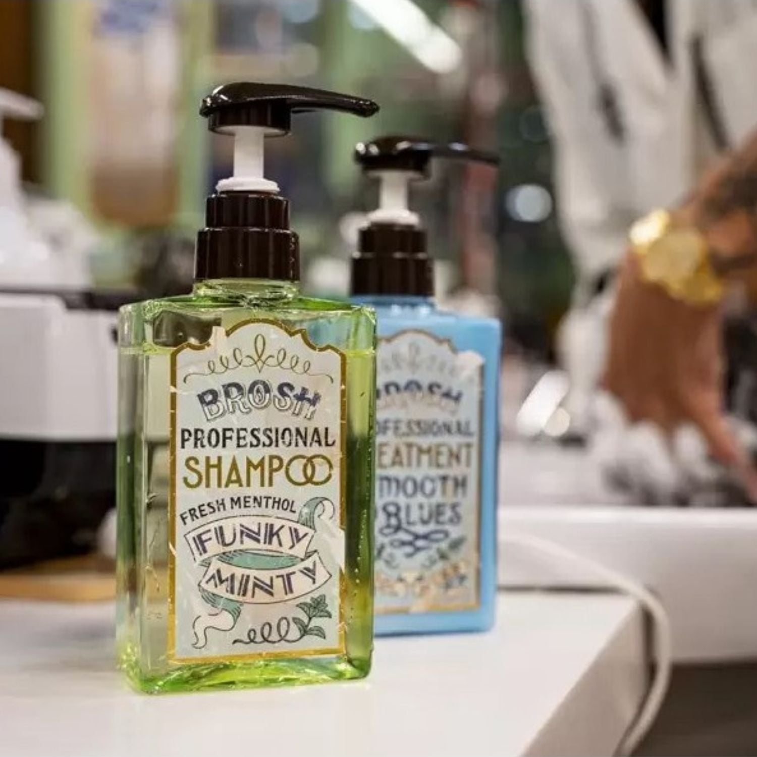 Two pump bottles of BROSH Professional Shampoo and Hair Treatment  on a white counter in a salon setting. The foreground bottle is clear and filled with a yellow-green liquid, labeled 'Fresh Menthol Funky Minty'.