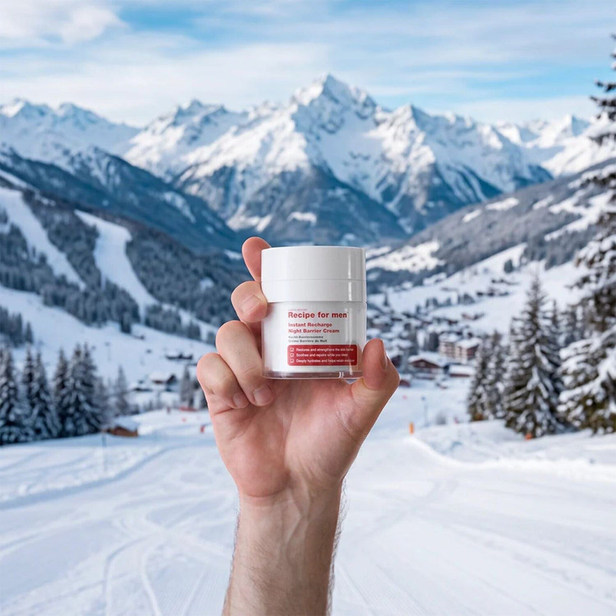 A hand holding a white cosmetic jar labeled 'Recipe for men' in front of a snowy mountain landscape.