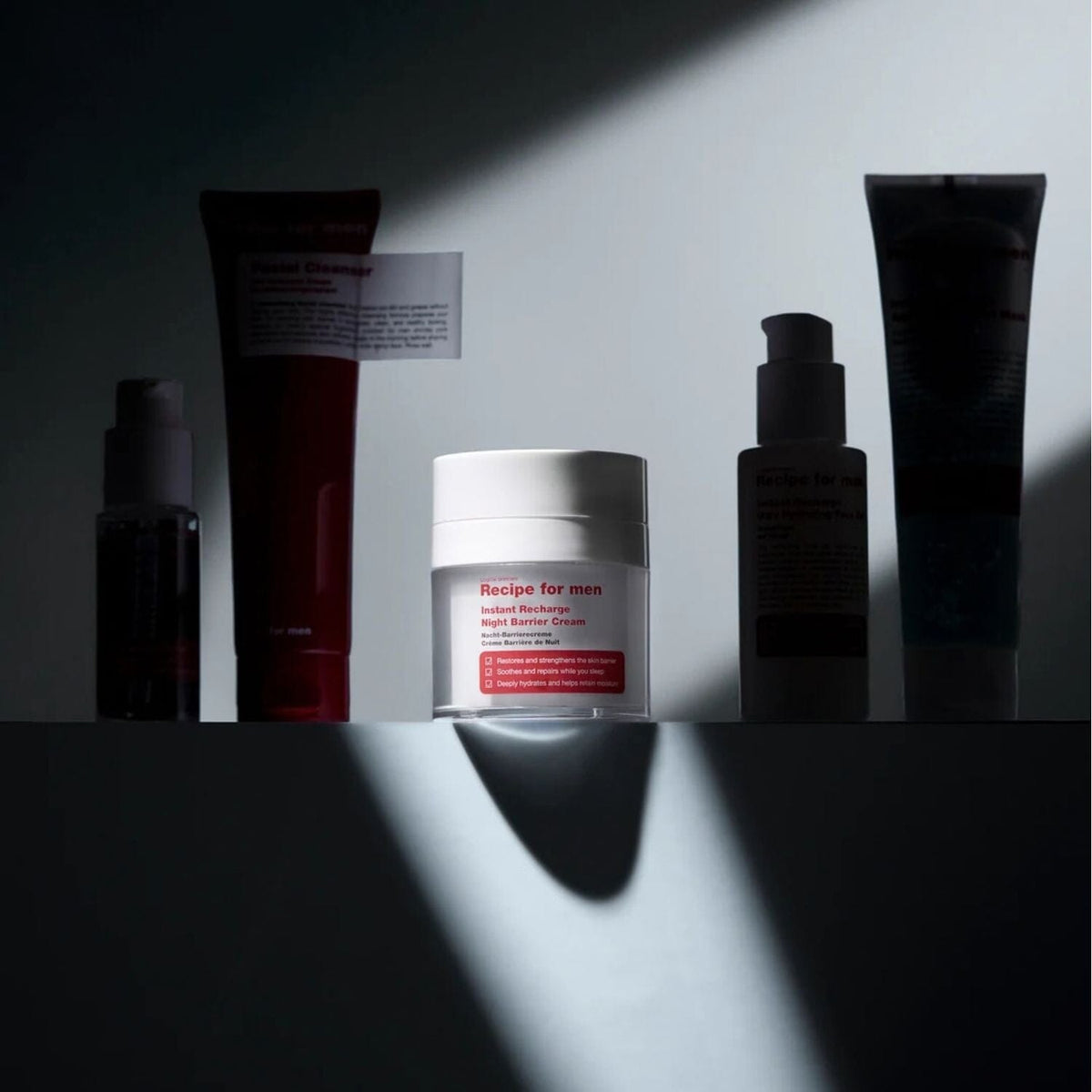 A lineup of skincare products from Recipe for Men on a dark surface under dramatic spotlighting. The lineup includes a dark bottle, a red tube labeled Facial Cleanser, a white jar labeled Instant Recharge Night Barrier cream, a dark bottle, and a tall dark tube.