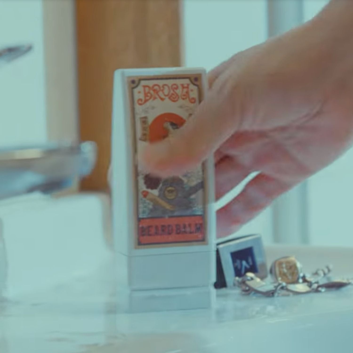A hand holds a small white rectangular product labeled 'BROSH BEARD BALM' on a light tabletop, with blurred bathroom-like background elements and a small card nearby.