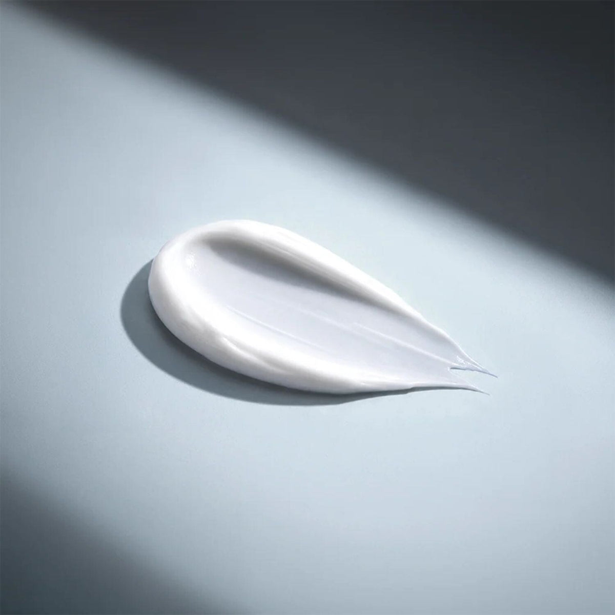 A smooth, creamy white product smear, likely a skincare cream or balm, displayed on a cool gray surface with dramatic diagonal lighting and soft shadows.