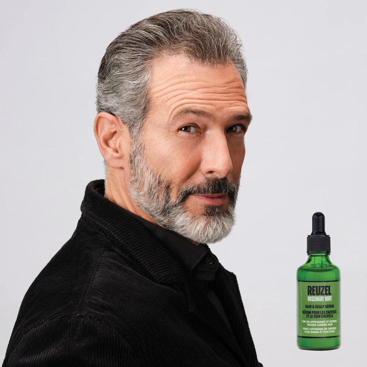 A man with gray hair and a gray beard wearing a black corduroy jacket, shown next to a green bottle of Reizel Hair Scalp Serum on a light gray background.