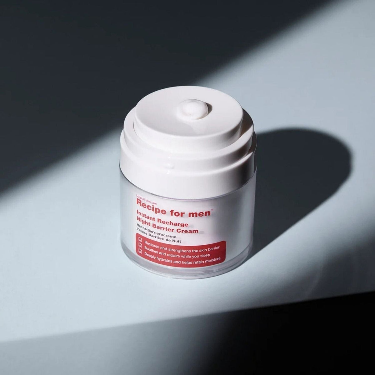 A clear cosmetic jar with a white screw-top lid sits on a light gray surface under sharp diagonal sunlight, casting a dark shadow. The label reads 'Recipe for men Instant Recharge Night Barrier Cream'.