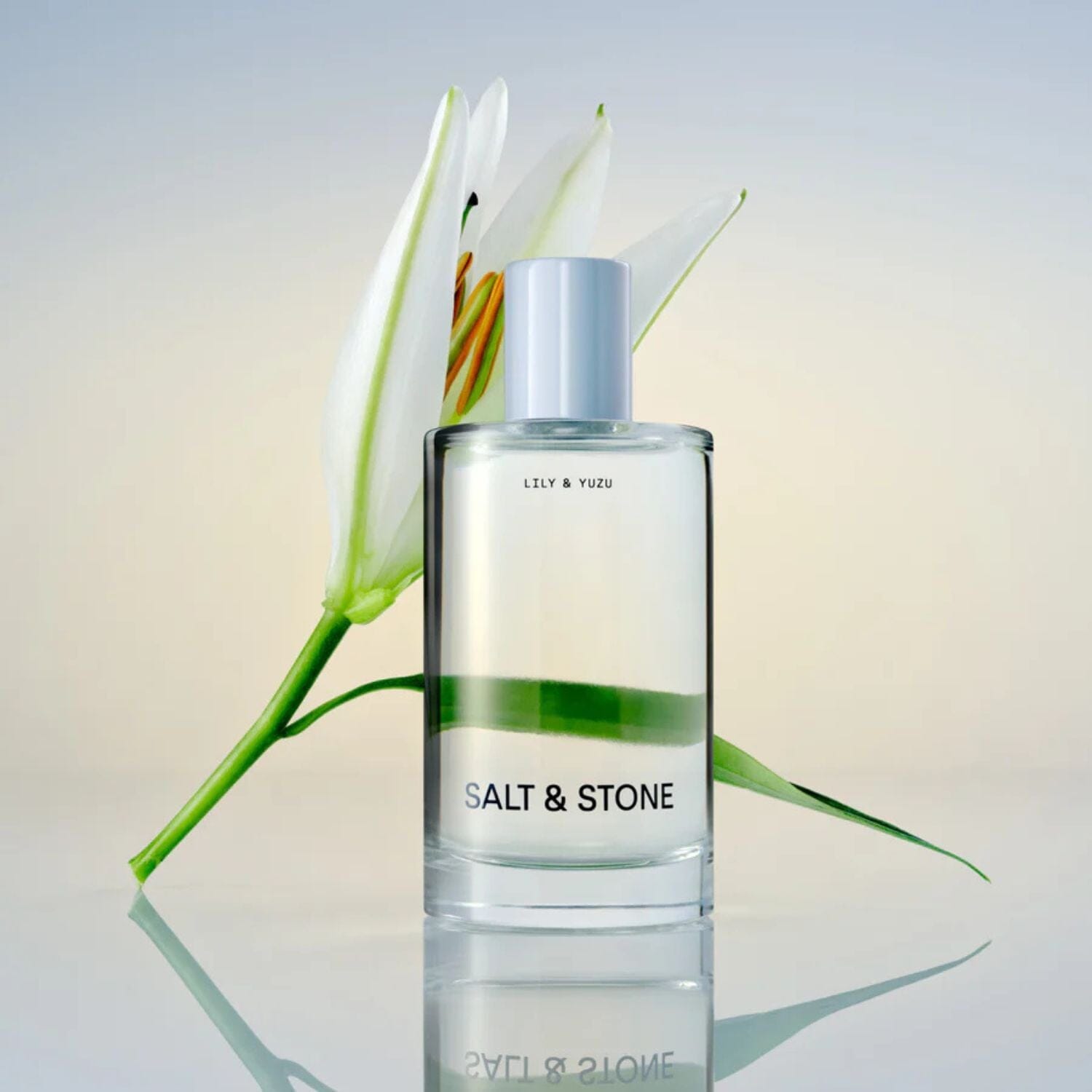Clear bottle of Lily & Yuzu Body Mist with a lily behind it on a reflective surface