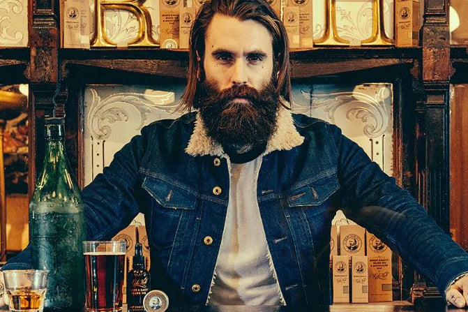 Man with a beard sitting at a bar with a bottle and glass in front of him.