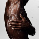 A close-up lifestyle shot of a man's bare torso and hand as soap lather is created on his skin against a light gray background.