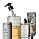 A collection of SALT & STONE skincare and body care products, including a pump bottle, two cylindrical jars, a spray bottle, and a squeeze tube, arranged against a plain white background.