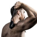 A shirtless man with a beard and short dark hair is shown from the back and side, holding a black handheld device against his head against a plain white background.