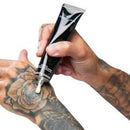 A close-up of tattooed hands holding a black squeeze tube and dispensing a white cream onto a surface against a plain white background.