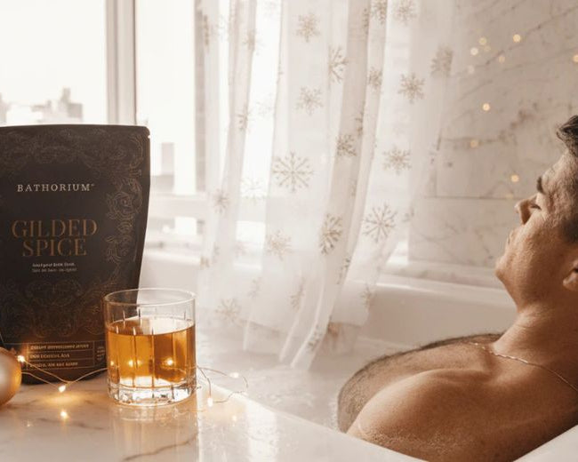 man soaking in a bath tub with the Gilded Spice Bath Soak Bag next to him and a beverage in a glass